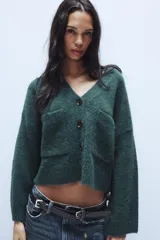 Chaqueta de punto de textura suave en color marrón oscuro, con escote en pico, manga larga y cierre frontal con botones. Presenta dos bolsillos de plastrón en la parte delantera.