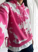 Buzo con capucha color fucsia y blanco con estampado tie-dye, cordones ajustables y lazos decorativos en los laterales.
