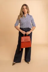 Mochila tipo cartera con tapa, confeccionada en símil cuero color suela. Cuenta con doble compartimento, división interna, cierre de seguridad y bolsillo trasero con cierre. Incluye una correa frontal decorativa con tachas metálicas.