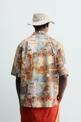 Camisa de corte relajado confeccionada en tejido ligero de algodón, con cuello bowling y manga corta. Presenta un estampado integral con motivos urbanos y tropicales en tonos cálidos.