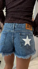 Short de jean azul con ruedo deshilachado, tiro medio, con una estrella blanca estampada en el bolsillo trasero derecho y etiqueta de cuero sintético con logo en la cintura.