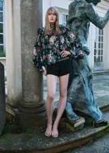 Blusa de gasa con estampado floral en tonos rosa, verde y negro. Tiene cuello alto, mangas largas con volantes y cierre de lazo en la parte posterior.