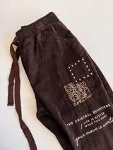 Pantalón de corte relajado color beige, con cintura elástica y cordón ajustable. Presenta parches decorativos con lentejuelas y tachas en los muslos, además de detalles de texto estampado. Posee bolsillos laterales y dobladillo en los tobillos.