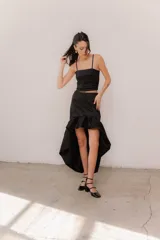 Pollera midi de tafeta color negro, con diseño asimétrico de corte mullet (más corta adelante y larga atrás), tiro alto y detalle de volado fruncido en el ruedo.