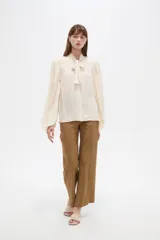 Camisa de poliéster color marfil, de diseño holgado, con cuello alto lazada y mangas largas abullonadas.