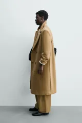 Abrigo largo color camel, de corte relajado (relaxed fit), confeccionado en tejido con mezcla de lana. Presenta cuello con solapas de muesca, manga larga, bolsillos de vivo en cadera, bolsillo interior y cierre frontal de botonadura cruzada.