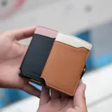 Billetera tarjetero automática con estructura de aluminio y revestimiento de cuero bicolor en tonos verde oliva y beige. Cuenta con un cierre de broche a presión y el logo de la marca grabado en la parte inferior.