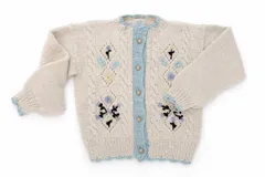 Cardigan de lana merino color marfil, con bordados florales artesanales, trenzas y rombos calados en el frente. Posee cierre frontal con botones vintage dorados y terminaciones en crochet celeste en cuello, puños, ruedo y tira de botones.
