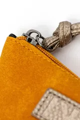 Billetera de gamuza color mostaza, con cierre superior y tirador de cuero sintético metalizado. Incluye placa metálica con grabado frontal.