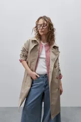 Trench clásico color marrón, de silueta relajada, confeccionado en tejido liviano, con cuello de solapa, doble abotonadura frontal y mangas largas con presillas regulables en los puños.