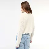 Chaqueta de punto con textura de croché en color beige, de corte corto y mangas largas. Presenta un cierre frontal mediante una lazada fina y bordes con acabado festoneado.