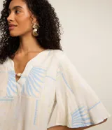 Blusa de manga corta en viscosa color beige, con escote en V con detalle de tira y cuenta de madera, y estampado geométrica y de follaje en tonos celestes.