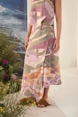 Falda midi confeccionada en lino con estampado camuflado en tonos rosado, verde y tostado. Presenta corte asimétrico con botones de madera frontales y detalles bordados en los ojales.
