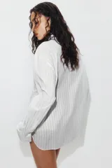 Camisa de popelín de algodón a rayas verticales, con cuello subido y aplicación de tejido tipo pañuelo anudado al frente. Presenta manga larga con puños anchos y cierre frontal con botones ocultos.