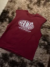 Musculosa sin mangas color bordó con cuello redondo. Estampado frontal en blanco con la palabra "HERMD" en un óvalo con efecto 3D y texto pequeño debajo que dice "el amor mueve el mundo y hasta el olimpo vibras del pasado. estilo del futuro".