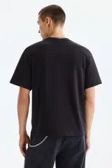 Remera negra de algodón con cuello redondo, manga corta y hombros caídos, de corte holgado. Presenta un estampado gráfico frontal estilo universitario en color blanco con efecto desgastado que lee "PROPERTY OF PHYS. ED. CLASS 1905 STATEN ISLAND".