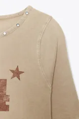 Camiseta color beige de algodón con efecto lavado, corte regular, cuello redondo con tachas metálicas, mangas cortas y estampado frontal con el número 74 y una estrella.