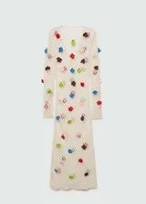 Vestido largo de tejido calado en color crudo, con diseño de red y aplicaciones de flores tejidas en diversos colores. Presenta escote en V con lazo, manga larga y corte entallado.