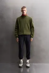 Chaqueta verde militar de punto con interior de polar. Tiene cuello alto, manga larga con bolsillo con cierre en la manga, bolsillos laterales con cierre y cierre frontal con cremallera.
