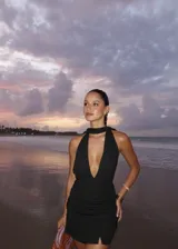 Vestido corto de tejido de punto con acabado brillante, escote profundo tipo halter y detalle de fruncido en la cintura. Presenta un diseño ajustado con una pequeña abertura frontal en el ruedo.