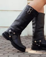 Bota de caña alta confeccionada en cuero negro, con suela track de goma. Presenta un diseño versátil con dos hebillas ajustables en la parte superior de la caña y una correa decorativa con tachas metálicas y argollas en la zona del tobillo.