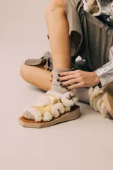 Sandalias de diseño moderno con tiras de cuero metalizado en color dorado, bordeadas con corderito sintético blanco. Poseen una suela plana con plataforma geométrica y cierre con hebilla lateral.