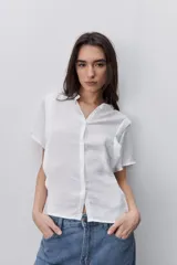 Camisa blanca de manga corta y silueta amplia, confeccionada en tejido liviano con textura sutil y caída fluida. Presenta cuello mao con pequeña abertura y cierre frontal con botones.