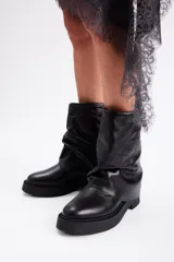 Bota de caña media confeccionada en cuero vacuno color marrón oscuro, con diseño de pliegues en la caña y suela de goma al tono con textura estriada.