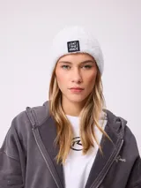 Gorro tipo beanie de punto acanalado en color gris claro, con parche rectangular negro en el frente que presenta el texto 'EAT THE WAVE' en color blanco.