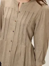 Camisa de lino mezcla color beige, con cuello redondo, cierre frontal con botones y mangas tres cuartos con puños elásticos. Presenta detalles de pespuntes verticales en zigzag en el frente.