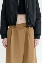 Pantalón sastrero de corte holgado en color tostado, con diseño asimétrico cruzado en la cintura, botón frontal y pliegues en las piernas.