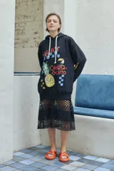 Buzo oversize de color negro con capucha y cordones ajustables. Presenta un estampado frontal con motivos tropicales, incluyendo una piña, un tucán y la inscripción 'Chiringuito'.