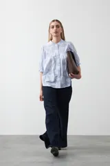 Camisa de algodón con fondo azul claro a rayas finas y estampa floral blanca en relieve. Presenta cuello cerrado, cartera de botones frontal y mangas cortas con volumen y puño elastizado. Posee frunces en el frente que aportan caída.