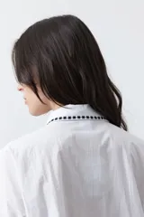 Camisa blanca de manga larga con detalles bordados en contraste a lo largo de la botonadura, el cuello y los puños. Presenta una silueta relajada y un diseño clásico con un toque artesanal.
