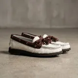 Mocasín de estilo náutico confeccionado en cuero, combinando una base de cuero con textura de pelo en color negro y detalles de cuero con textura croco en color marrón. Presenta cordones decorativos con borlas laterales y suela plana de goma.
