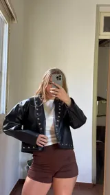 Campera corta de ecocuero negro, con diseño de solapas rectas decoradas con tachas metálicas plateadas a lo largo de los bordes frontales y el ruedo. Presenta mangas largas de corte amplio y estilo moderno.