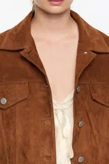 Cazadora corta de corte oversize confeccionada en piel caprina con acabado de ante. Presenta cuello camisero, cierre frontal con botones metálicos, bolsillos con tapeta en el pecho y bolsillos de vivo en el bajo. Incluye mangas largas con pliegues en el codo y puños abotonados.