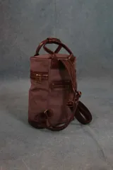 Mochila matera de diseño vertical, confeccionada en tela canvas marrón con detalles y refuerzos en cuero sintético oscuro. Cuenta con doble asa superior de mano, correas traseras regulables, cierre metálico frontal y bolsillo posterior.