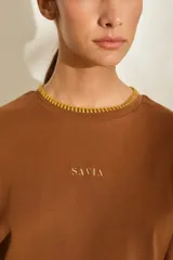 Remera marrón de manga corta y cuello redondo, con detalles de costuras decorativas en contraste color crudo en el cuello, los puños y el dobladillo inferior. Cuenta con el nombre de la marca impreso en el pecho.