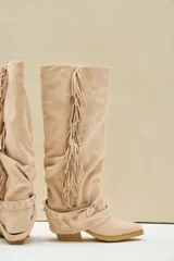 Bota texana alta de gamuza color beige, con taco cuadrado, flecos largos colgantes en el lateral y tiras con hebilla metálica alrededor del tobillo.