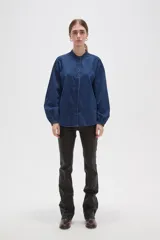 Camisa de jean azul con cuello mao, cierre frontal con botones y mangas largas con puños elásticos fruncidos.