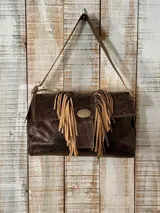 Cartera tipo bandolera de cuero marrón con efecto desgastado, solapa frontal decorada con flecos beige y placa metálica central, y asa larga ajustable.