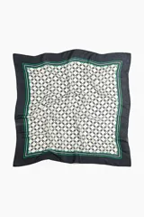 Pañuelo cuadrado de tejido suave con estampado geométrico central en tonos crema y negro, enmarcado por bordes negros con líneas decorativas en verde y celeste.