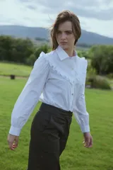 Camisa blanca de corte clásico con cuello camisero, mangas largas abullonadas y detalle de volados en el canesú delantero. Presenta cierre frontal con botones.