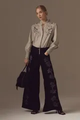 Blusa de algodón con estampado de rayas finas, cuello alto con volados y cierre de cordón. Presenta bordados florales en el pecho y mangas largas con puños elásticos.