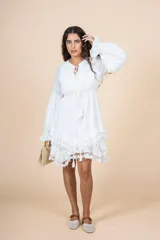 Vestido corto color blanco, de corte holgado con cintura elástica. Presenta escote en V con lazo de ajuste, mangas largas abullonadas con puños elásticos y falda con volados decorados con encaje en el ruedo.