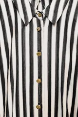 Vestido midi combinado con parte superior de camisa a rayas verticales blancas y negras, cuello solapa, manga sisa con frunces y botones dorados. La parte inferior es una falda midi negra de corte recto. Incluye un cinturón negro con textura de cocodrilo y hebilla dorada.