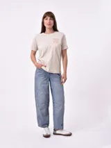 Remera de manga corta color beige claro con cuello redondo. Presenta un estampado gráfico central con un plato de pasta rodeado por un borde azul y blanco, sobre un fondo de franjas verticales rojas y blancas. El estampado incluye texto en cursiva en tonos turquesa y rojo.