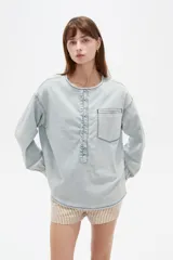 Blusa de jean ligera en color celeste claro, con cuello redondo y cierre frontal parcial con botones. Presenta un diseño con volados a lo largo de la botonera, bolsillo ojal en el pecho, y mangas largas abullonadas con puños ajustados y fruncidos.