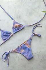 Conjunto de bikini con corpiño triangular de atar con apliques de argollitas y bombacha vedetina de atar con apliques de argollitas. El tejido presenta un estampado tie-dye o marmolado en tonos lila/azul y naranja/beige.
